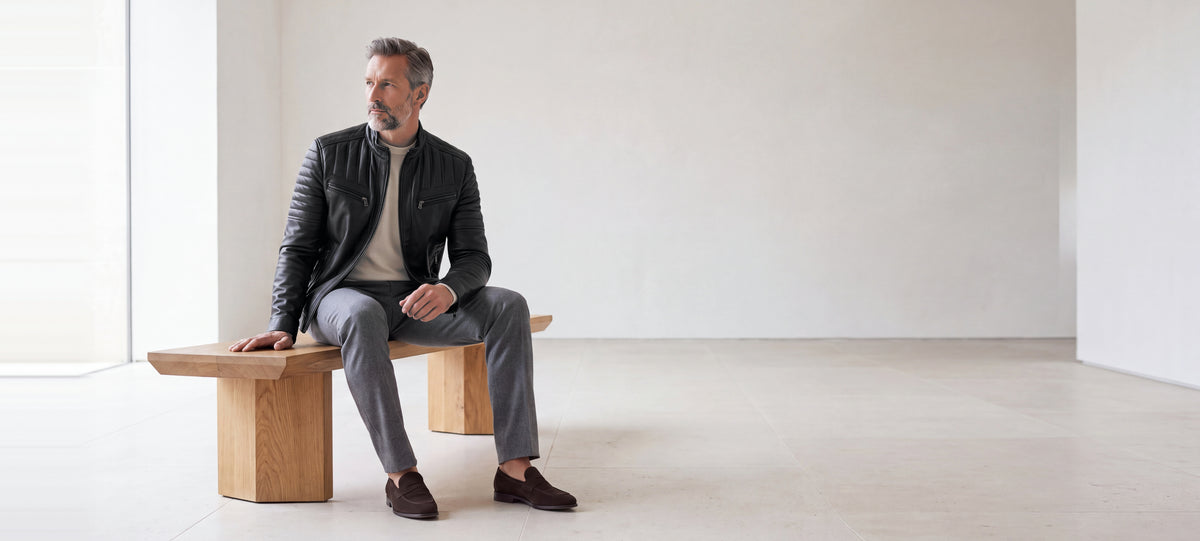 Racer Varenne en cuir d'agneau matelassé noir porté, assis sur un banc en chêne dans un intérieur minimaliste